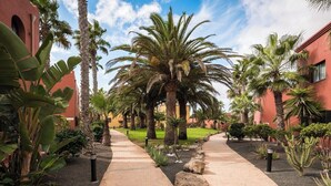 A pathway flanked by palm trees and a building with a red facade. - Home2Book Oasis Paradise Home Fuerteventura (Corralejo)