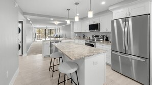 Une cuisine moderne dotée d’un grand îlot central, d’appareils électroménagers en acier inoxydable et de luminaires suspendus.