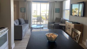 Una sala de estar con sofá, sillas, una mesa de comedor con un tazón de cereal, un televisor y un balcón con vistas.