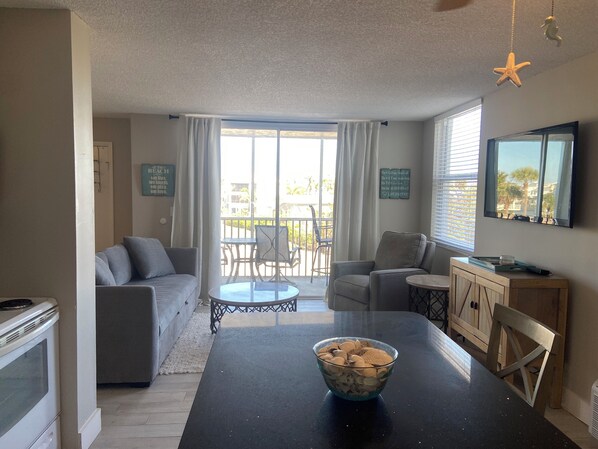 A living room with a sofa, chairs, a dining table with a bowl of cereal, a TV, and a balcony with a view.