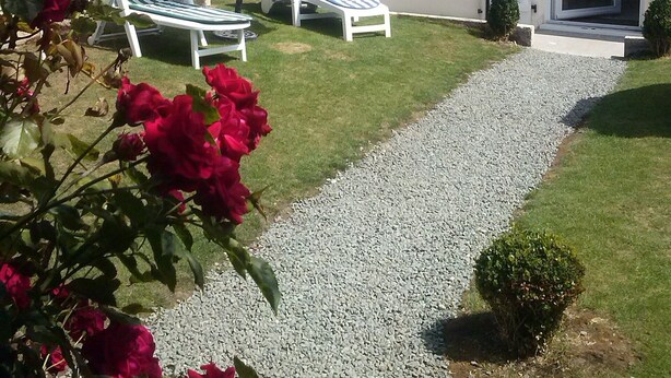 Een huis met een grindpad dat naar een witte deur leidt, twee witte lounge fauteuils onder een blauwe parasol, en rode rozen op de voorgrond.