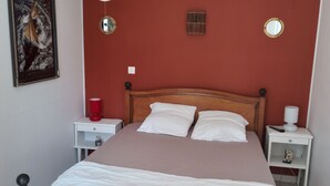 Een net opgemaakt bed met wit en bruin beddengoed, een houten hoofdbord met decoratieve metalen details, en een rode muur met twee wandspiegels en een klein plankje met een lamp en een afstandsbediening.
