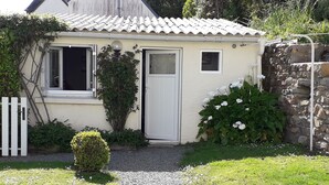 Een klein, laag huis met een pannendak, een witte deur en een raam. Een grindpad leidt naar de deur, omringd door gras en planten.