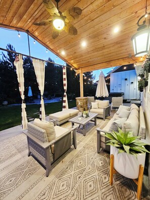Un patio exterior con techo de madera, asientos acolchados, alfombra estampada y una planta colgante.