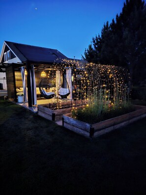 Una casa con porche cubierto, un jardín con bancales y luces colgantes.