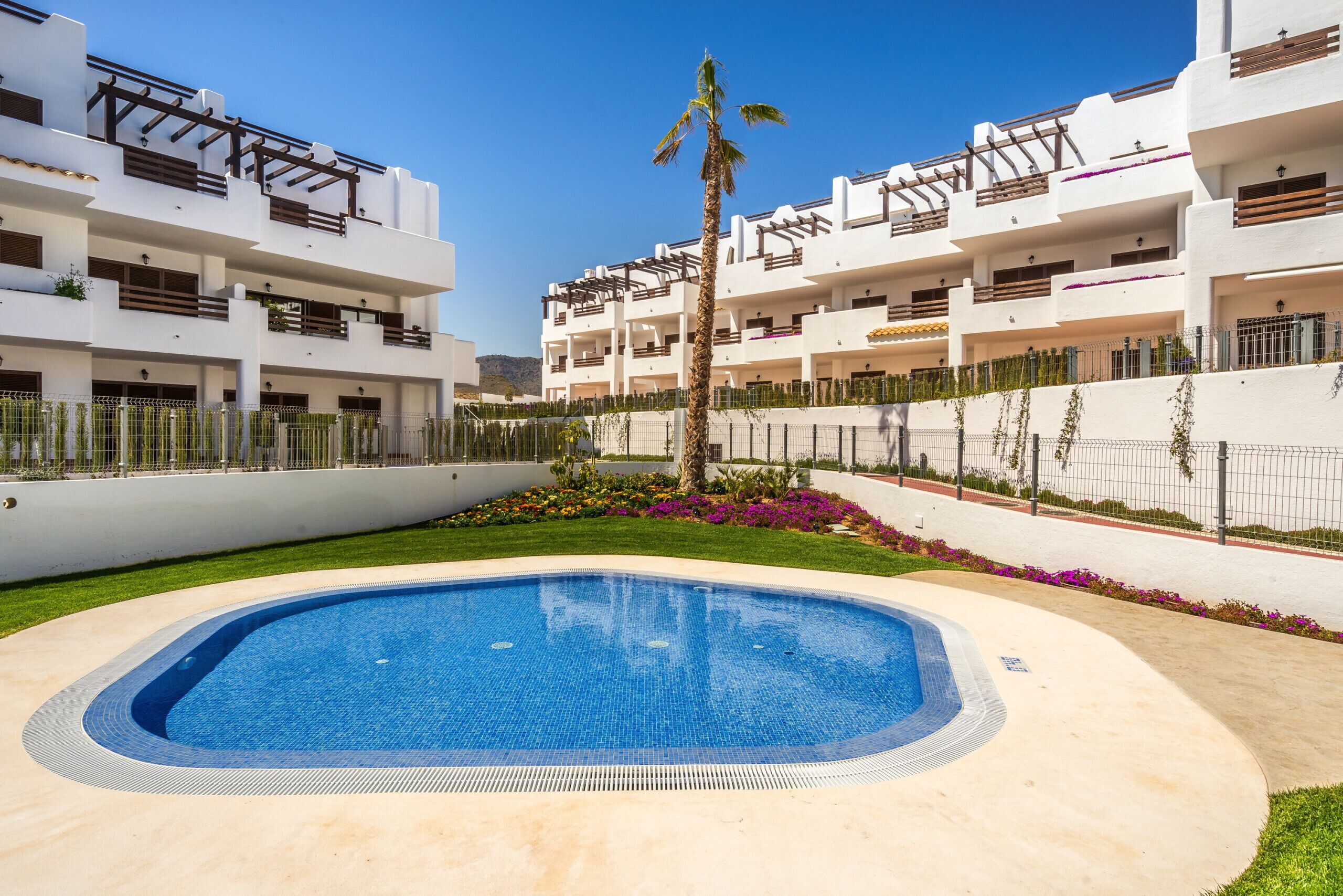 A modern building with a swimming pool surrounded by a well-maintained garden and a clear blue sky.
