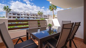 Ein Balkon mit Glastisch und Stühlen, Blick auf einen Pool und ein Gebäude mit Palmen.