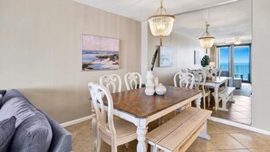 A dining area with a wooden table and white chairs, a chandelier, and artwork on the walls. - Windward Pointe 604 | Spacious, Update, Beach Front! (Orange Beach)