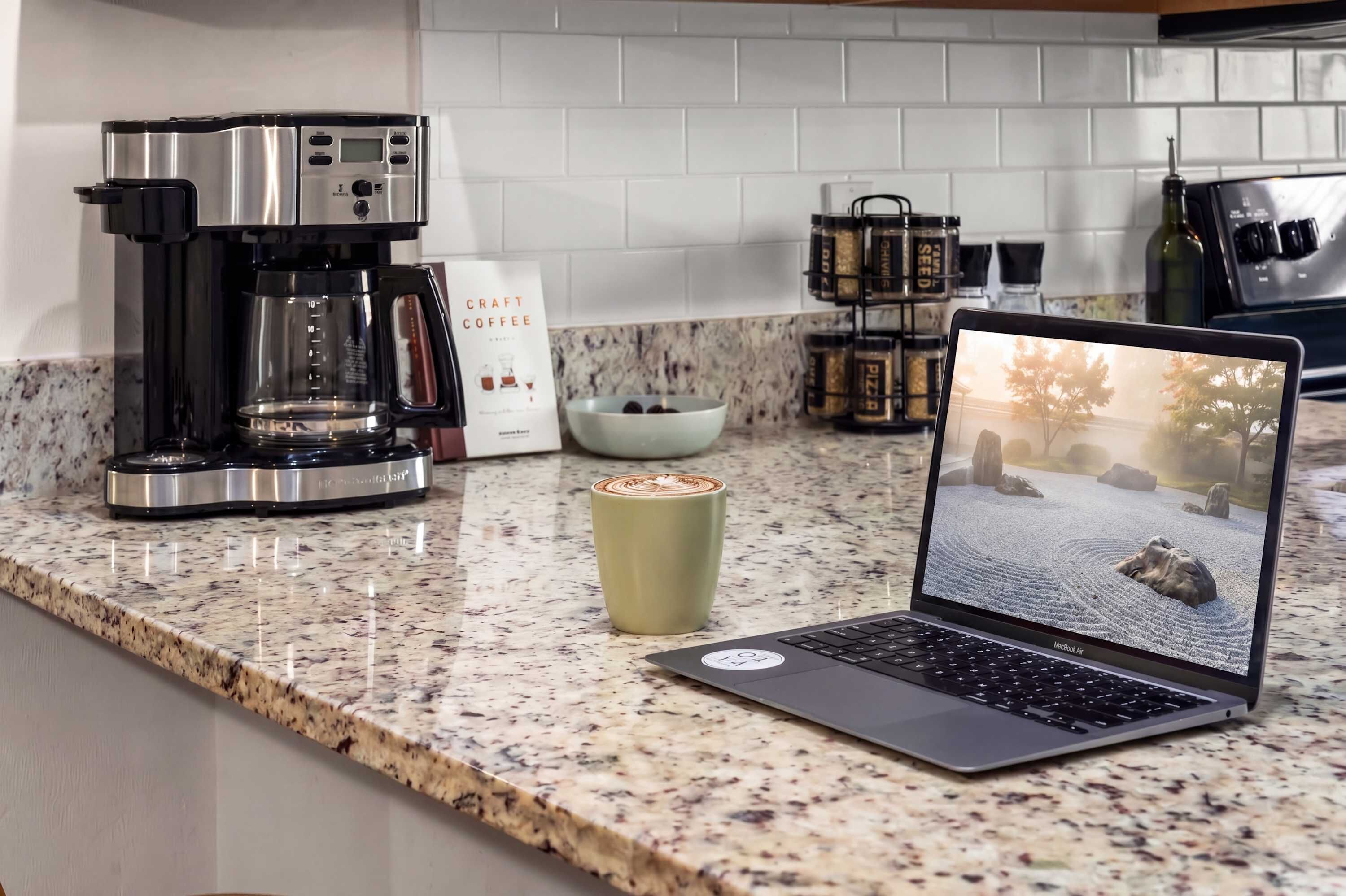 En køkkenbordplade med en kaffemaskine, en bærbar computer der viser et naturmotiv, en kop og en kaffepose.
