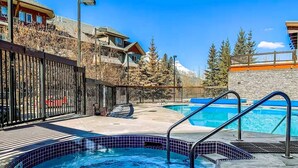 A swimming pool area with a hot tub, a wooden deck, and a view of a mountain. - 🥇Renovated 2BR, AC, Heated Pool, Hot Tub, Pets ok! (Canmore)