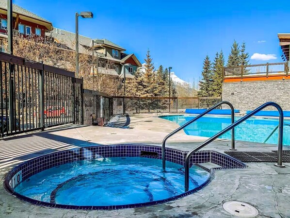 A swimming pool area with a hot tub, a wooden deck, and a view of a mountain.