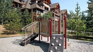 A playground with a climbing structure and a slide, surrounded by trees and a lodge in the background. - 🥇Renovated 2BR, AC, Heated Pool, Hot Tub, Pets ok! (Canmore)