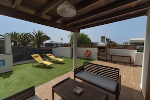 A covered patio area with a wooden bench, yellow lounge chairs, and a table.