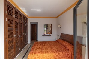 A bedroom with a patterned bedspread, wooden wardrobes, a mirror, and a bedside lamp.