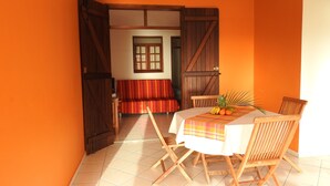 A dining area with a table set for two, a wooden bench, and a door leading to another room. - GITES + SWIMMING POOL AND SPA, 5 Mn FROM MALENDURE BEACH AND VIEW OF THE PIGEON ISLANDS   (Bouillante)