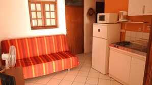 A compact kitchen with a refrigerator, microwave, and stove. A striped sofa is positioned against the wall, and a fan is on the floor. - GITES + SWIMMING POOL AND SPA, 5 Mn FROM MALENDURE BEACH AND VIEW OF THE PIGEON ISLANDS   (Bouillante)