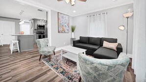 En moderne stue med en mørkegrå hjørnesofa, et hvidt sofabord og to møbelstole med et mønster. Loftsventilator og indbygningsspots er synlige.