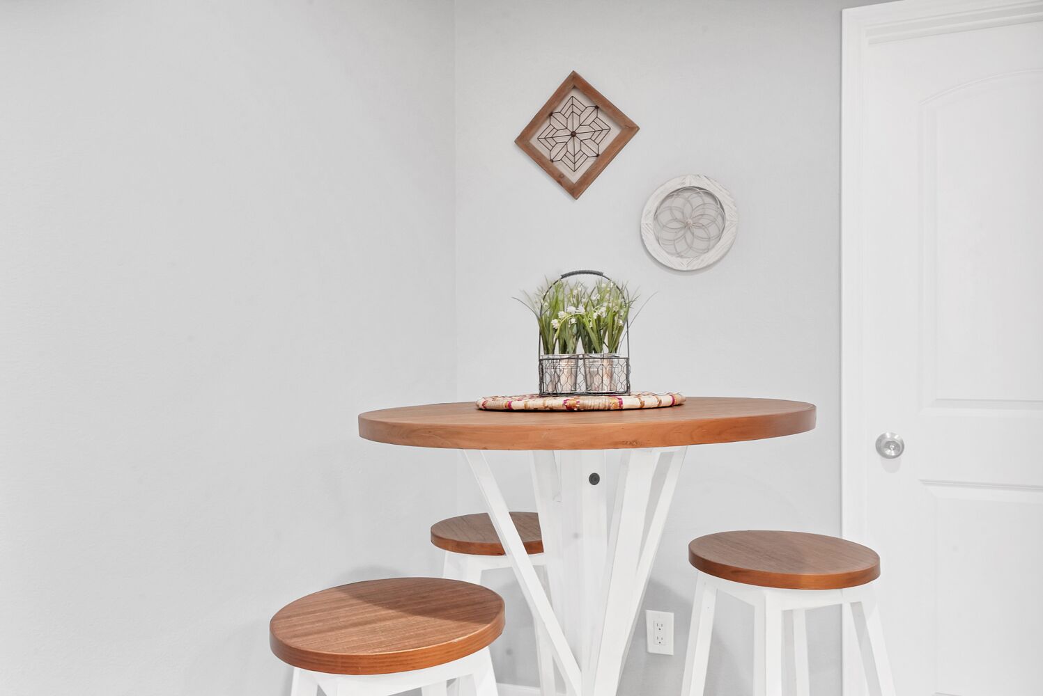 A round wooden table with a woven placemat and a metal basket with green plants. There are three white stools with wooden tops.