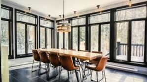 A dining area with a long wooden table and six chairs, large windows, and a chandelier. - Chalet cheri (Mont-Tremblant)