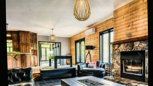 A cozy living room with a stone fireplace, wooden walls, and a modern fireplace insert. There is a leather sofa, a coffee table, and a dining table with chairs. - Chalet cheri (Mont-Tremblant)