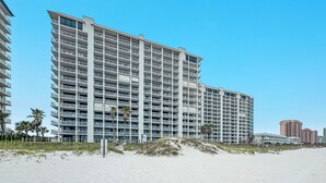 A high-rise hotel with multiple balconies, located near a beach with sand dunes and palm trees.