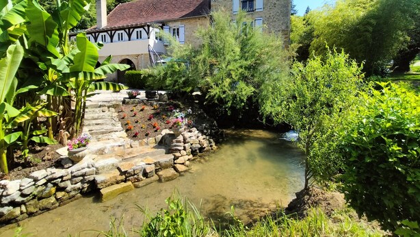 Ein Haus aus Stein mit einem steilen Dach, umgeben von üppigem Grün und einem kleinen Bach.