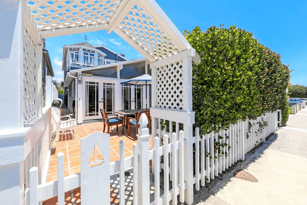 A white gazebo with a lattice roof, a patio area with a table and chairs, and a neatly trimmed hedge along a white picket fence.