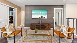 Una sala de estar con un televisor grande montado en la pared, un mueble de madera debajo y dos sillones de cuero con cojines.