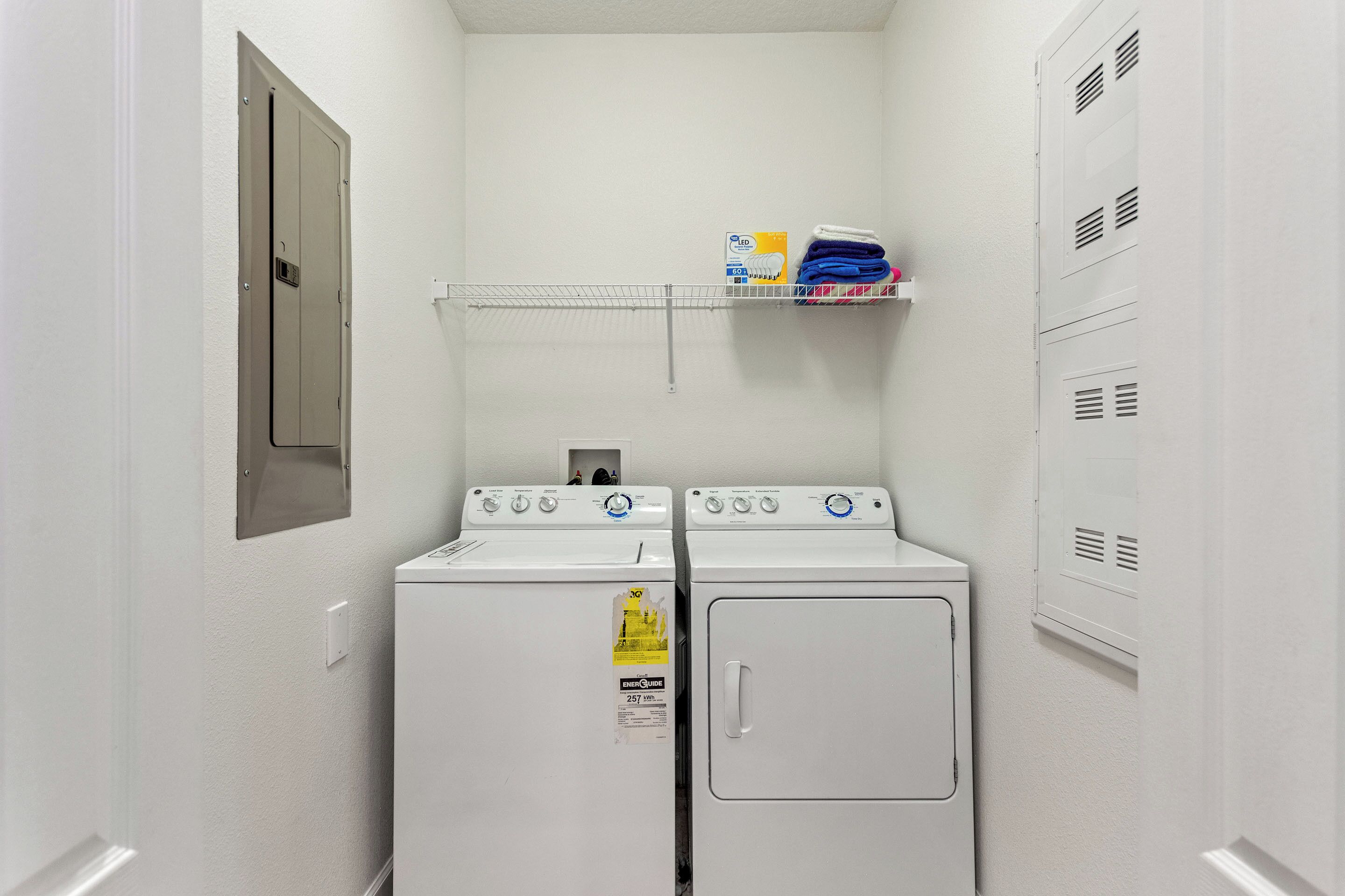 A laundry room with two front-loading washers and dryers, a shelf with laundry supplies, and a door with a security camera.