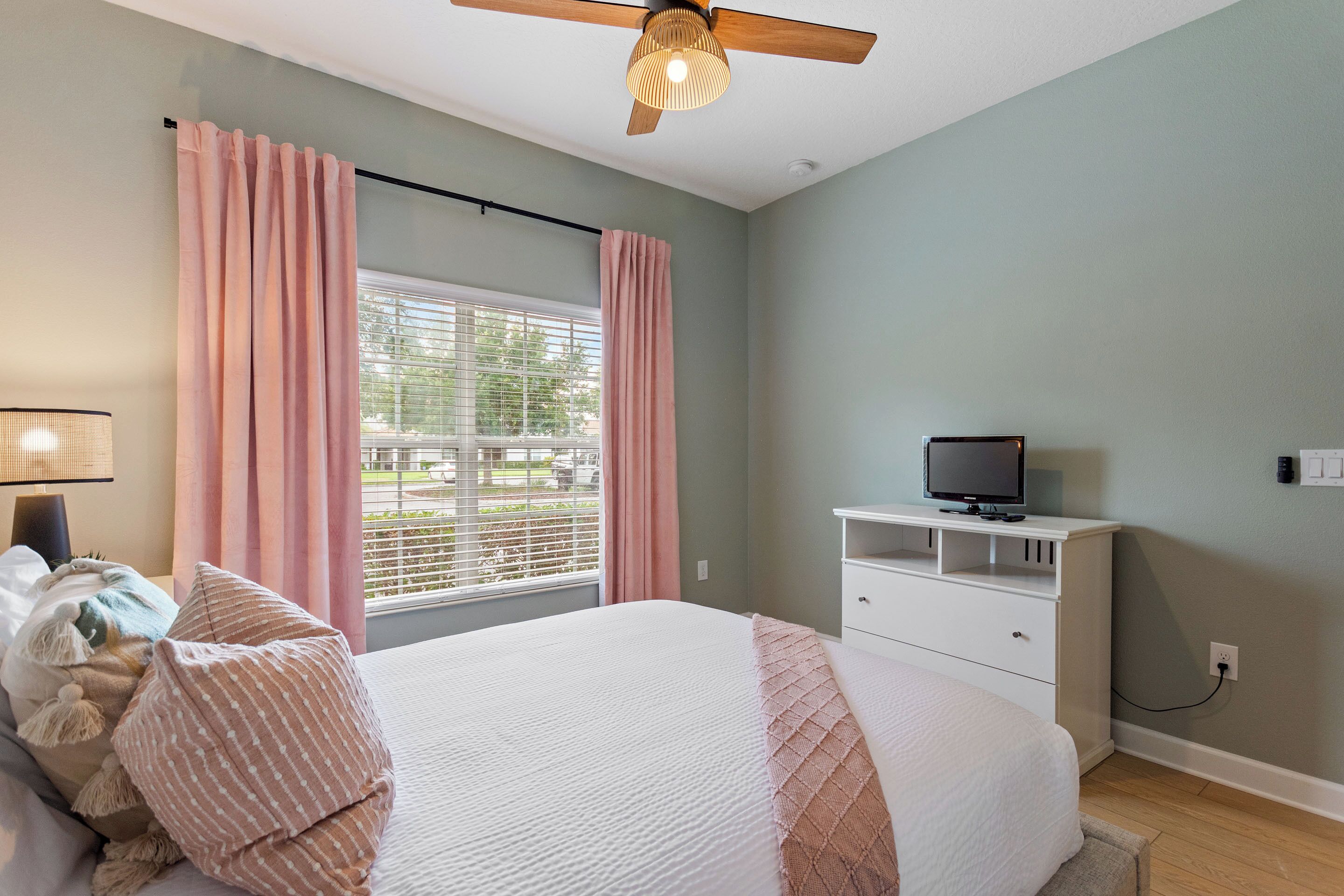 Une chambre à coucher avec un lit, un téléviseur posé sur un meuble, une fenêtre avec des rideaux et un ventilateur de plafond.