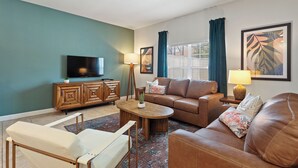 A living room with a brown sofa, a white armchair, a wooden coffee table, a TV on a wooden cabinet, and a window with curtains. - Private Pool, Premium Resort Amenities Included (Kissimmee)