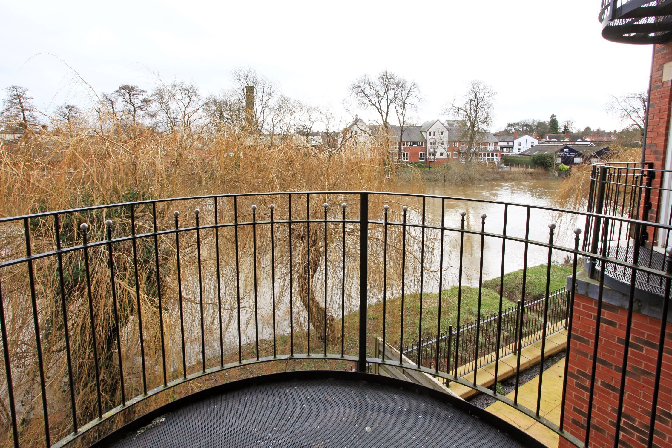 A view from a balcony overlooking a river with a metal railing and a brick building in the background.