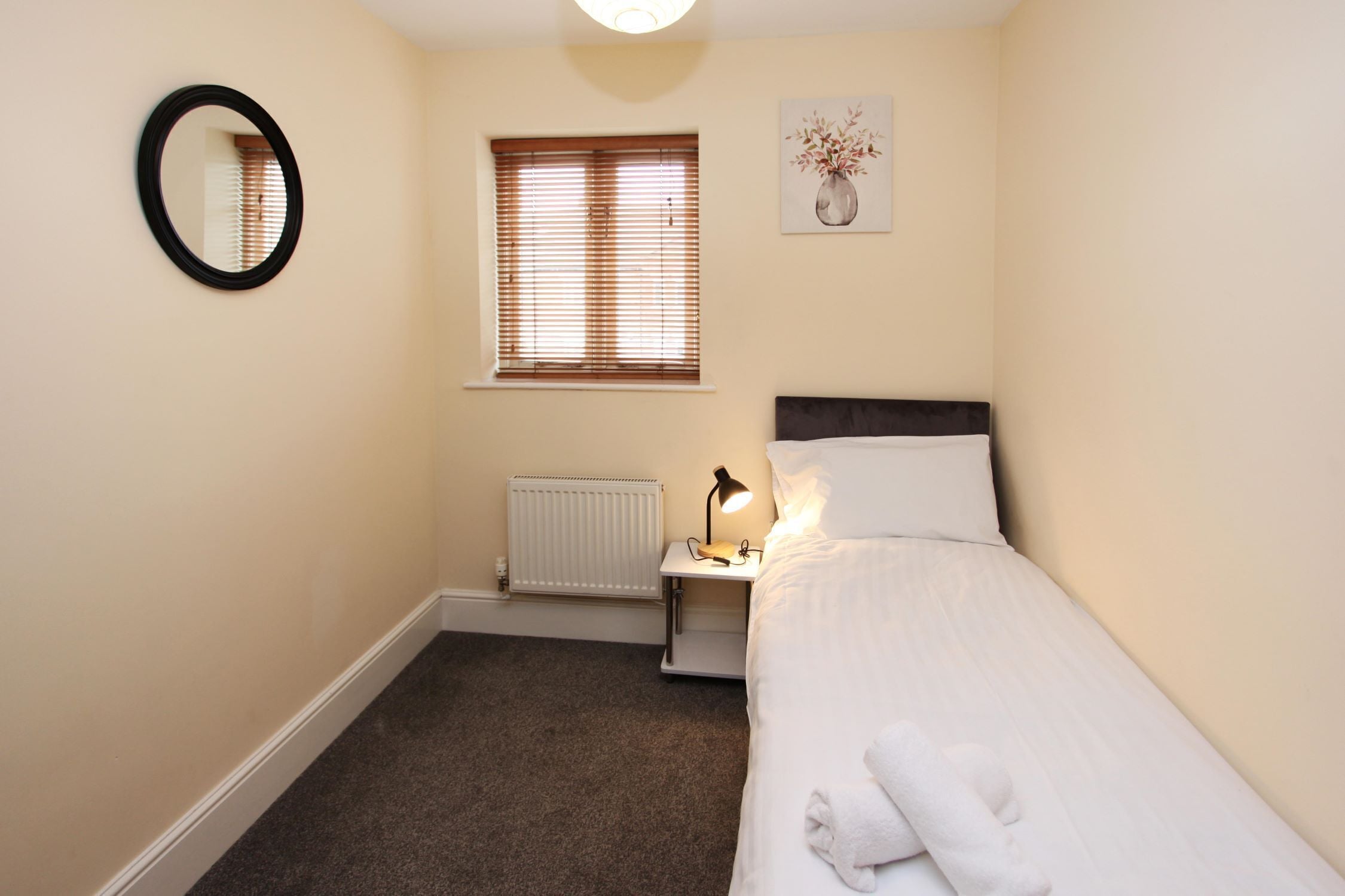 Een slaapkamer met een bed, een nachtkastje met een lamp, een radiator, een raam met jaloezieën, een spiegel aan de muur en een schilderij.