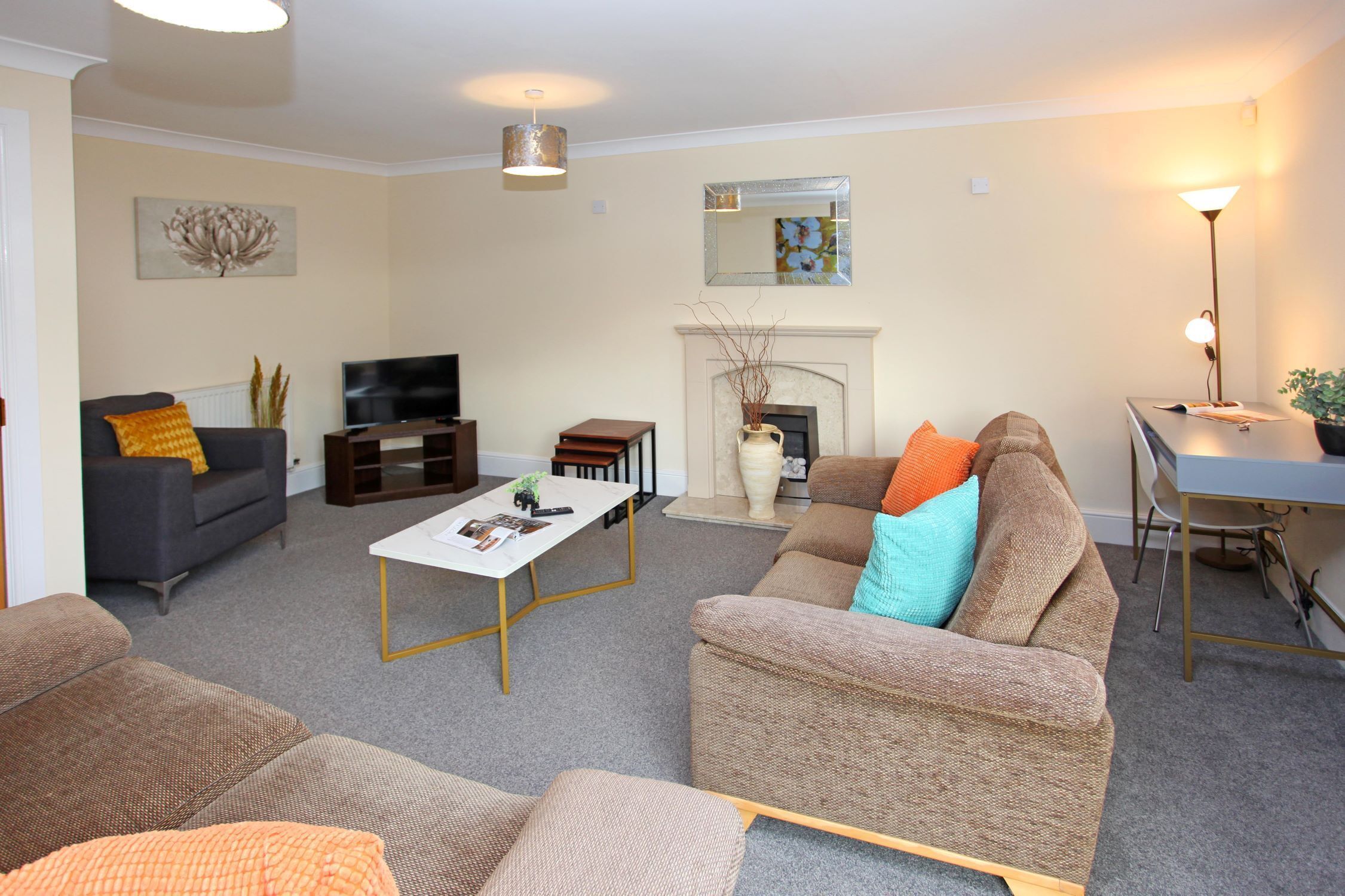 A living room with a sofa, armchairs, a coffee table, a fireplace, a TV, and a desk with a lamp.