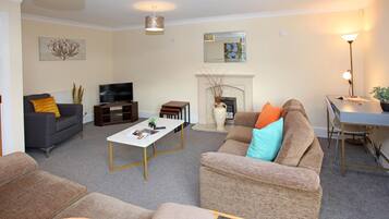 A living room with a sofa, armchairs, a coffee table, a fireplace, a TV, and a desk with a lamp.