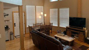 A spacious living room with a high ceiling, wooden beams, a large window, a television, a sofa, a dining table, and a kitchen area. - Cozy Cabin with Modern Amenities (Pet Friendly) (Pagosa Springs)