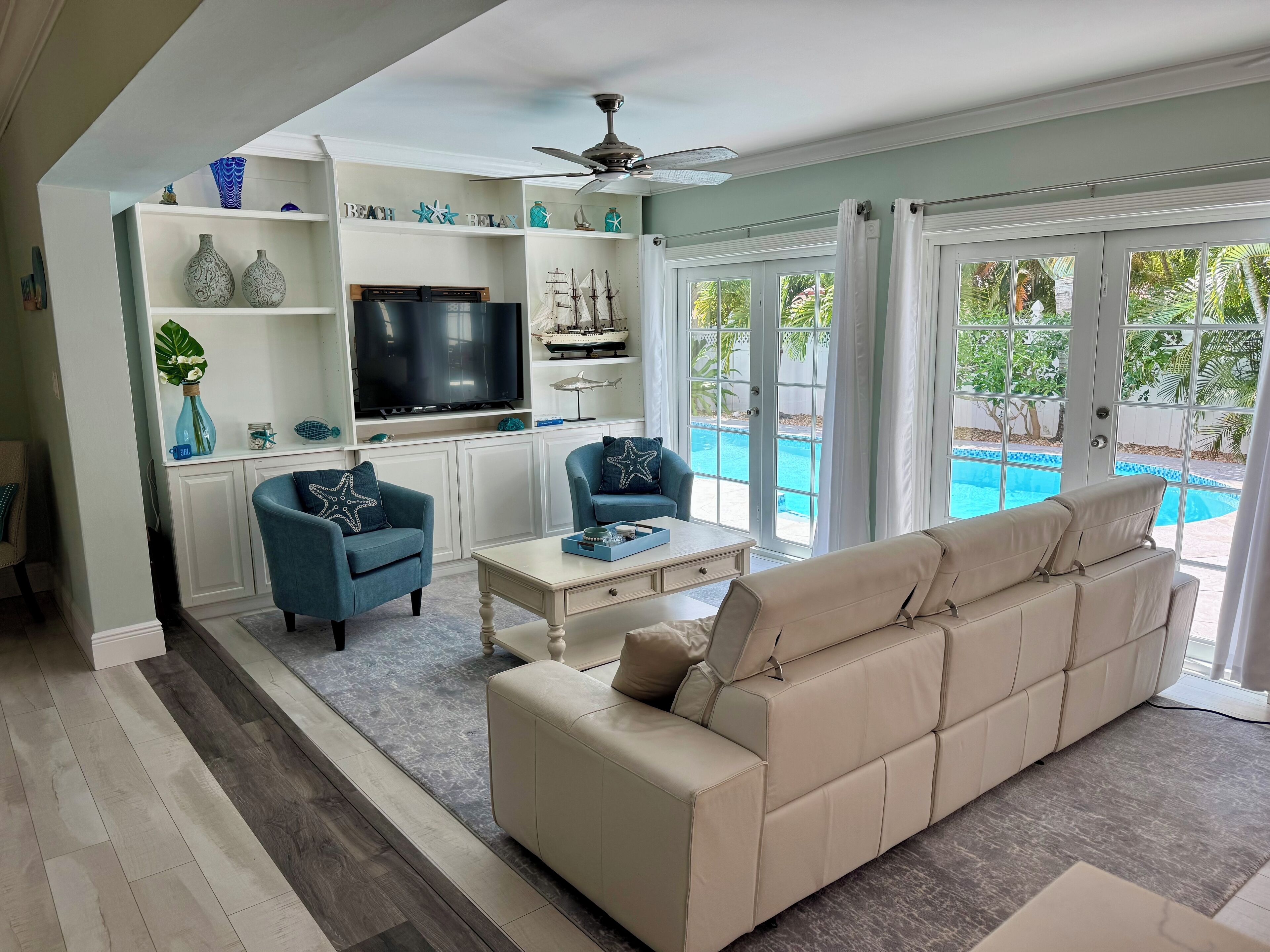 A living room with a beige sofa, a blue armchair, a coffee table, and a TV. There is a pool visible through the glass doors.