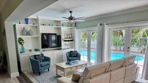 Una sala de estar con un sofá beige, un sillón azul, una mesa de centro y un televisor. Se ve una piscina a través de puertas de vidrio.