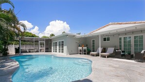 Una piscina con un deck de piedra, un patio cubierto con asientos, y una casa con ventanas grandes.