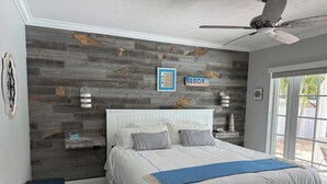 Un dormitorio con una cama blanca, un ventilador de techo, un panel de pared de madera y una ventana con vistas a la vegetación.