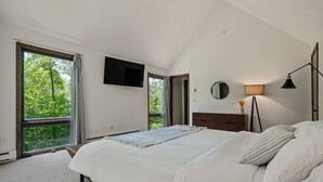 Un dormitorio con una cama grande, un televisor en la pared, una lámpara de noche y vistas a árboles a través de las ventanas.