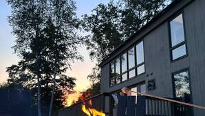 Una chimenea con llamas, rodeada de mobiliario exterior, frente a un edificio con grandes ventanales y una terraza de madera.