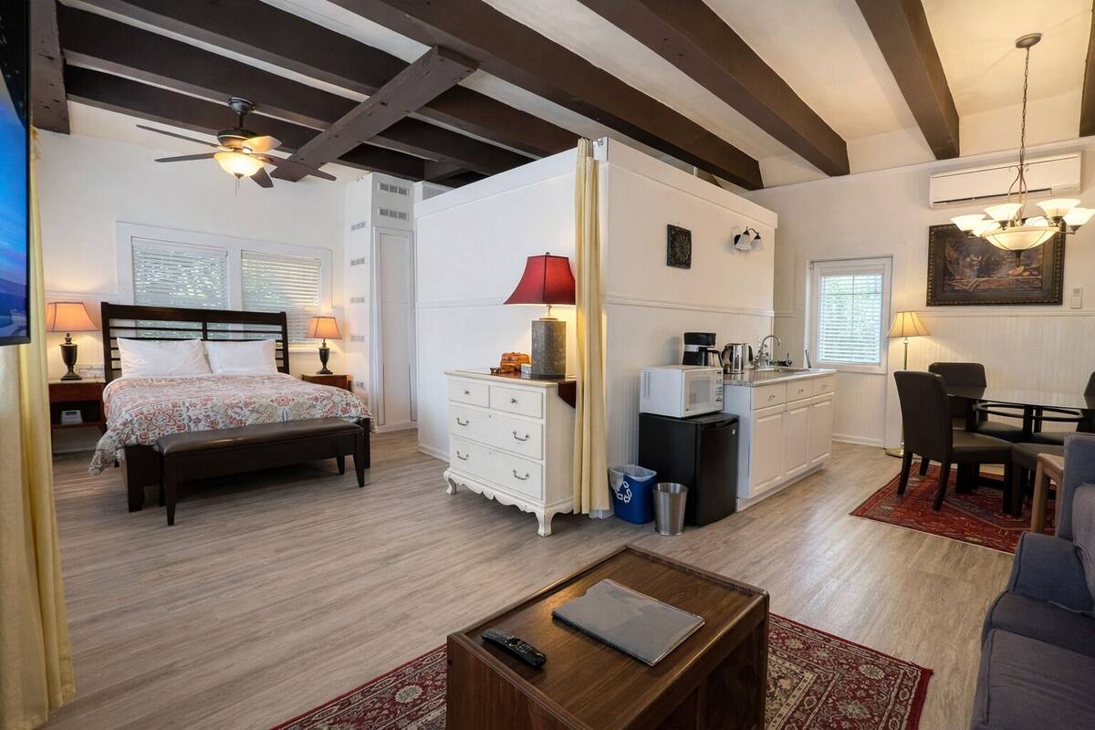 Un dormitorio con cama, mesitas de noche, escritorio, silla, kitchenette pequeño y zona de comedor con mesa y sillas.