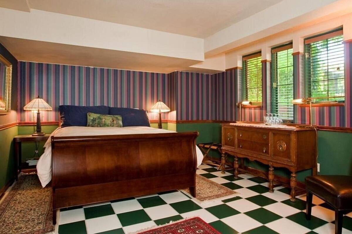 Un dormitorio con cama de madera, paredes verdes, papel tapiz a rayas, suelo de cuadros, cómoda de madera y dos lámparas.