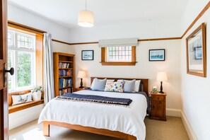 Ein Schlafzimmer mit einem großen Bett, einem Holzregal, zwei Nachttischen mit Lampen, einem Fenster mit Vorhängen und gerahmten Bildern an der Wand.