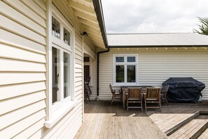 Eine Holzterrasse mit Tisch und Stühlen, einem Grill und einem Haus mit weißer Außenverkleidung und einem Metalldach.