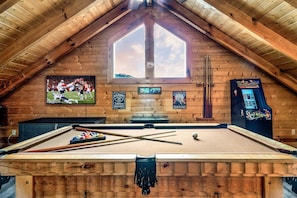 A pool table in a wooden room with a large window, a mounted TV, and a whiskey sign.