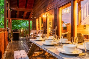 Een houten terras met eettafels, buitenverlichting en uitzicht op de omliggende begroeiing.