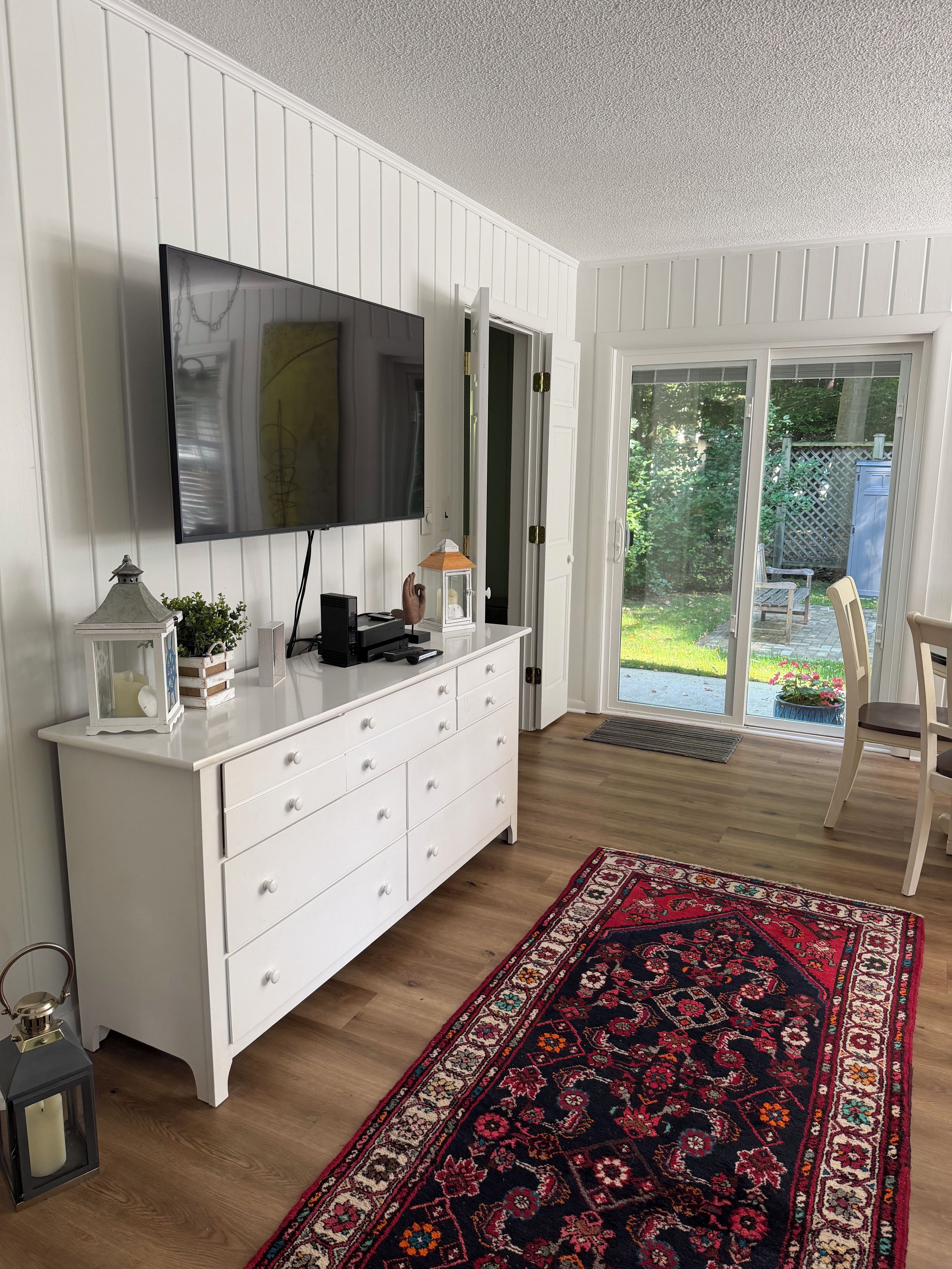 A room with a white dresser, a flat-screen TV, a patterned rug, and a sliding glass door leading to an outdoor area.