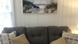 Un sofá gris con cojines estampados, un cuadro de una playa y una ventana con persianas.